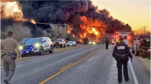 Image of a large fire started by an 18-wheeler and train collision with public safety personal parked and standing in the street