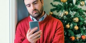 Image of a sad man in a red sweater looking at his phone in front of a Christmas tree