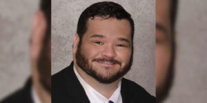 Image of Man with dark hair and beard wearing a black suit, black tie, and white button up shirt