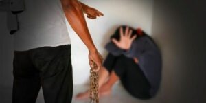 Image of man holding a chain pointing at a scared woman in a corner who is covering her face and holding up her hand