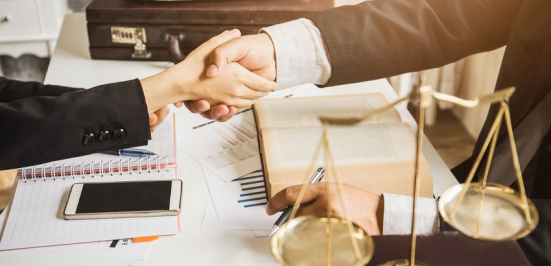 Image of two people shaking hands over a desk covered in paperwork next to a legal scale