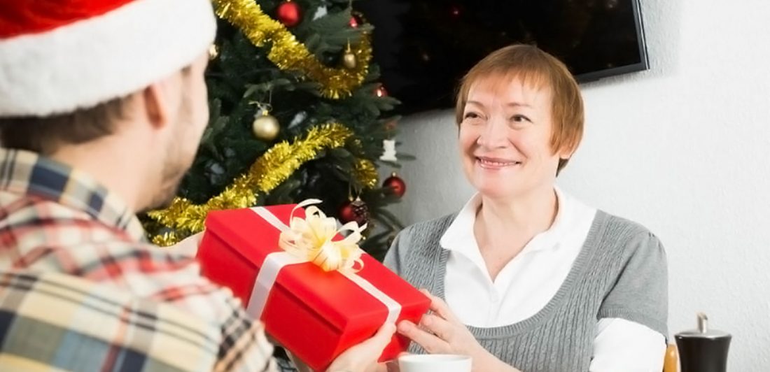 Image of older woman giving a red gift with a cream colored bow to a man in a santa hat in front of a Christmas tree