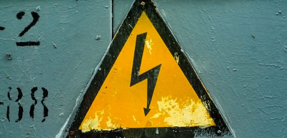 Image of a yellow and black triangle sign with a lightning bolt with an arrow on the end on a green wall